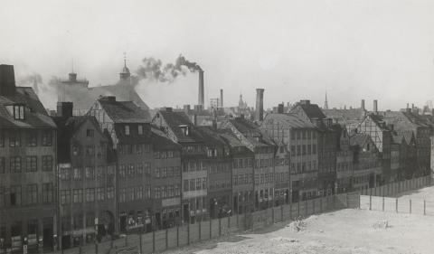 Den sorte røg fra de tidlige badeanstalters kuldrevne anlæg betød, at naboerne ikke kunne have vinduer åbne, uden at fint kulstøv lagde sig over det hele. Her er det røgen fra den hambroske badeanstalt ved Trinitatis Kirke. I forgrunden ses den ryddede plads efter det bortsanerede Brøndstrædekvarter, der stødte op til Vognmagergade. Billedet er fra 1911 (foto: Københavns Museum).